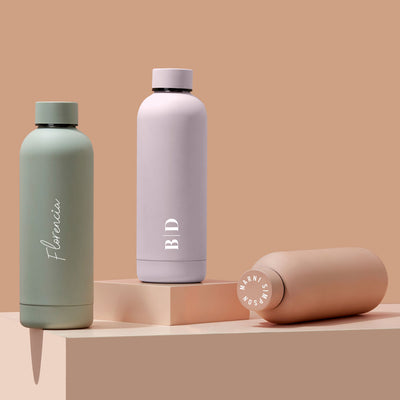 Three insulated stainless steel water bottles with different colors (green, white, and pink) and a beige base. The green bottle has the name 'Flora', the white has 'BD', and the pink has a logo or design.